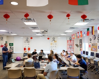 A classroom with TCU students seated at desks, each with laptops and notebooks. A person stands at the front near a display screen. The room is decorated with international flags and red lanterns. The walls have educational posters and various artworks.