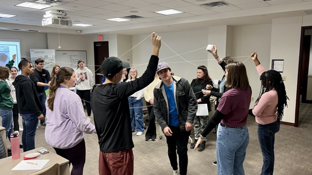Students standing in a circle holding string above their heads. 