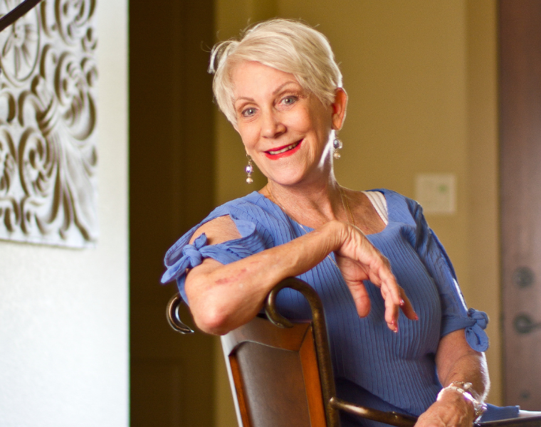 Beverly Bass sitting on a chair, wearing a blue top with ribbon accents on the sleeves, smiles warmly. The background features a decorative wall piece.