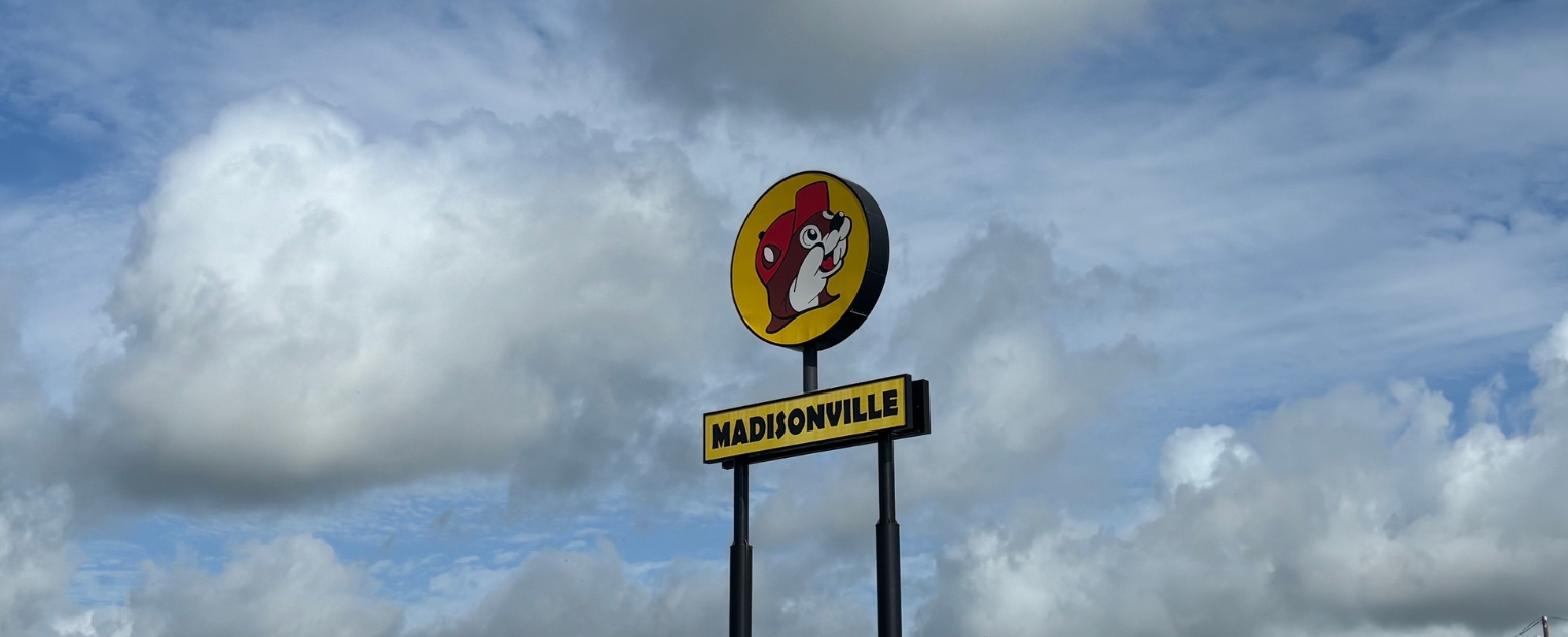 A person in a Buc-ee's beaver costume wearing a red cap and shirt stands in the store. The logo "Buc-ee's" featuring a beaver is visible on the wall and on merchandise.