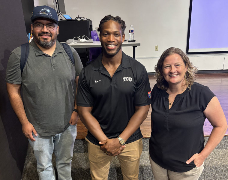 Three smiling individuals stand together indoors. The person in the center is wearing a TCU shirt. The person on the left is wearing a cap and carrying a backpack. The setting appears to be a classroom or lecture hall with a screen in the background.