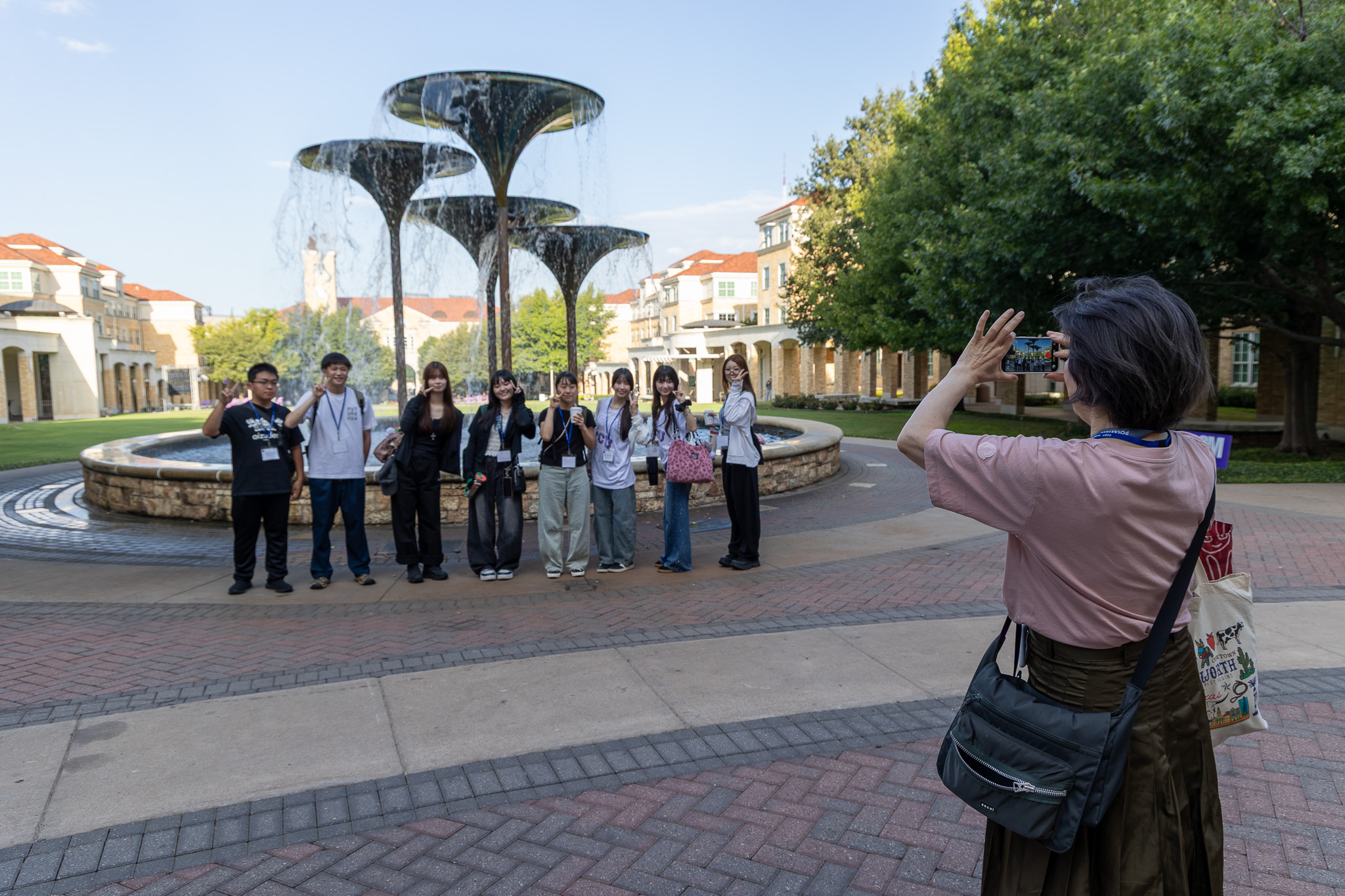 A group of students from Japan poses for a photo in front of Frog Fountain. Another person stands to the side, capturing the scene with a smartphone. 