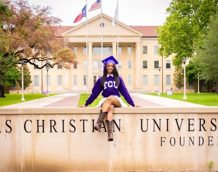 Jaya Armstead in a graduation cap and TCU sweater sits on a wall in front of Texas Christian University, with the building and flags visible in the background.
