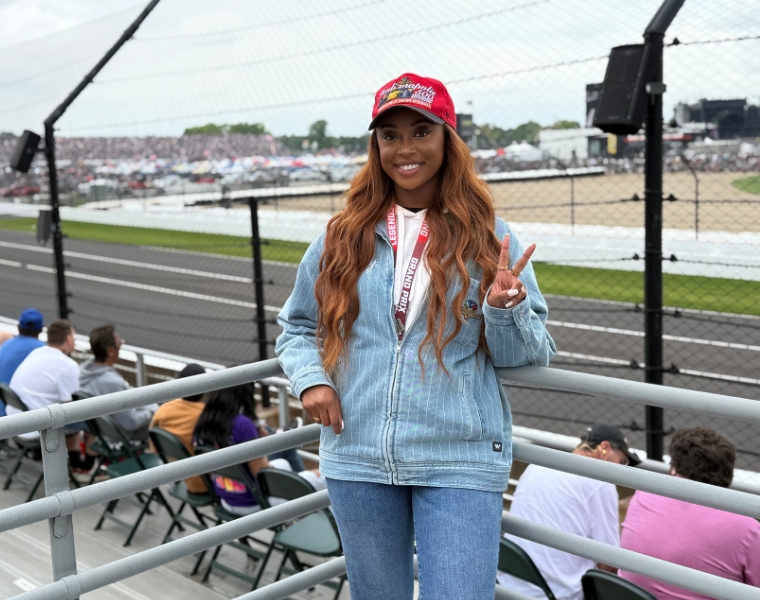 Jaya Armstead stands by the fence at a car racing track, wearing a denim jacket, jeans, a red Indy 500 cap, and a lanyard with a pass. She is smiling and making a peace sign gesture. The racetrack and spectators are visible in the background.