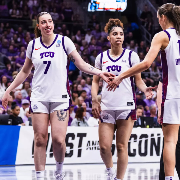 A group of TCU basketball players on the court during a game, wearing white jerseys with purple accents. The background shows an audience and banners, indicating a lively game atmosphere.