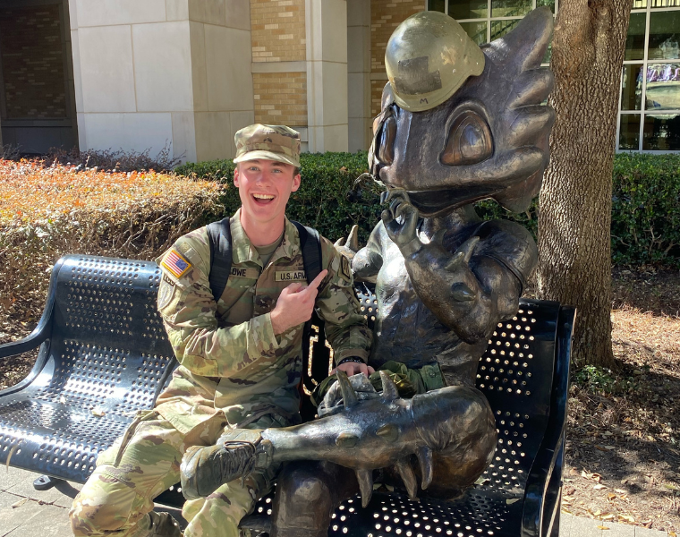 Matt Lowe in military uniform is sitting on a bench beside a statue of SuperFrog wearing a helmet. The statue is making a Go Frogs gesture. The environment appears to be outdoors with bushes and a building in the background.
