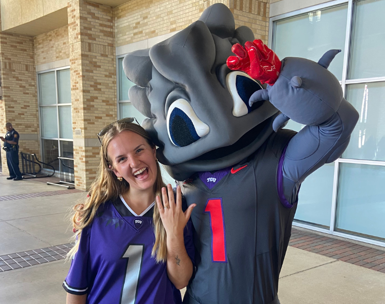 A person wearing a purple TCU jersey poses happily next to the TCU mascot, SuperFrog, who is in a playful stance. They are standing outside near a building with large windows.