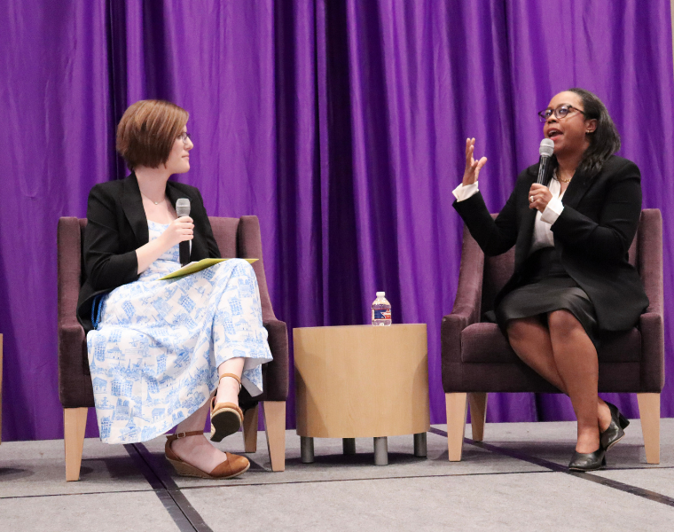 Two individuals are seated on a stage with microphones, engaging in a discussion. One is wearing glasses and gesturing with a hand while speaking. A small table with a water bottle separates them. The background features purple curtains.