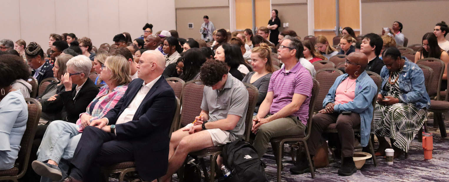 Two individuals are seated on a stage with microphones, engaging in a discussion. One is wearing glasses and gesturing with a hand while speaking. A small table with a water bottle separates them. The background features purple curtains.