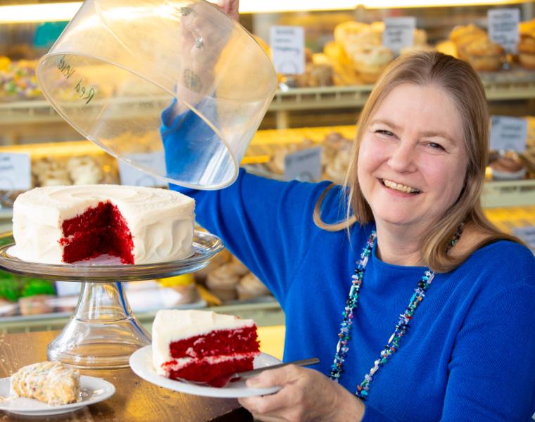 Rebecca Sharpless smiling while lifting a cake cover and holding a slice of red velvet cake.