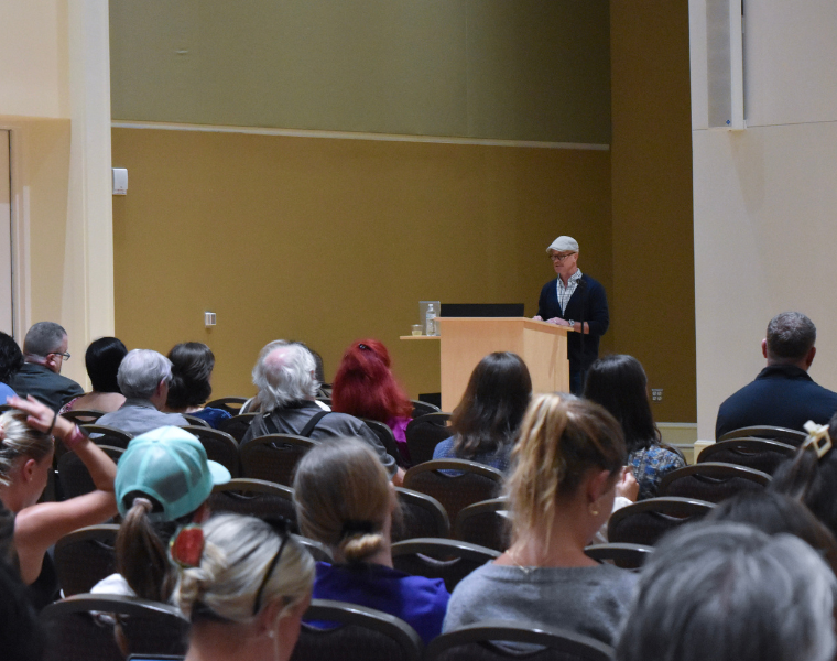 Sage Elwell stands at a podium speaking to an audience seated in an auditorium. The audience is facing the speaker, and some individuals are taking notes.