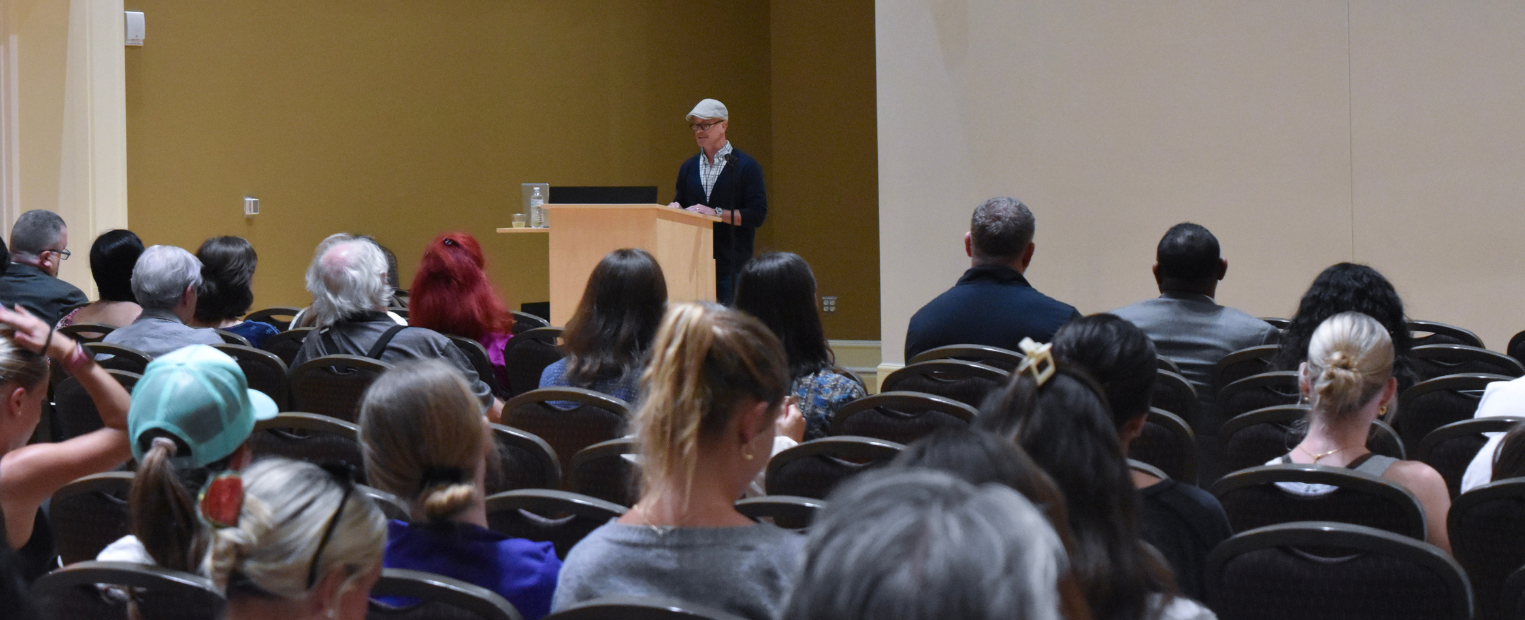Sage Elwell stands at a podium speaking to an audience seated in an auditorium. The audience is facing the speaker, and some individuals are taking notes.