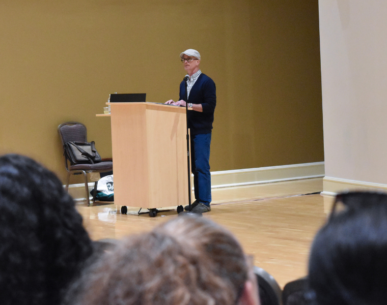 An individual wearing a hat and glasses speaks at a wooden podium in a lecture hall. The background is a plain mustard-colored wall, and there are some seated audience members visible in the foreground.