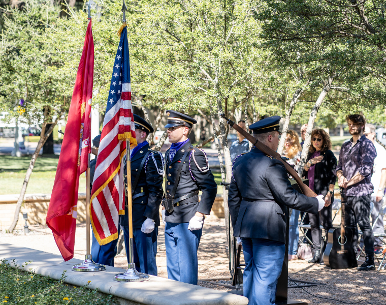 A group of people stand near a pair of flags, including the United States flag, in a park setting. Some individuals are dressed in military uniforms, holding a rifle and a bugle. Others are standing nearby, watching the scene. Trees and sunlight create a calm outdoor atmosphere.