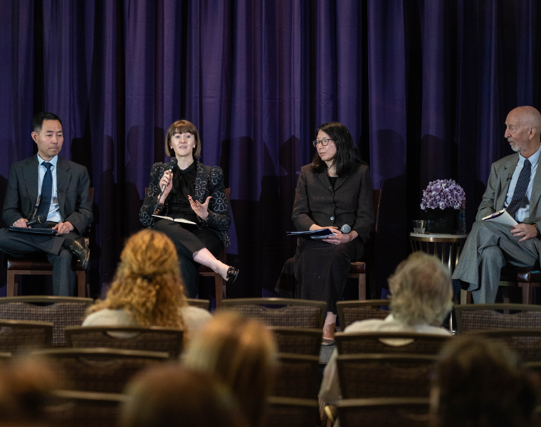 A group of four people are seated on a stage in front of a purple curtain, participating in a panel discussion. Two individuals hold microphones. Audience members are visible in the foreground.
